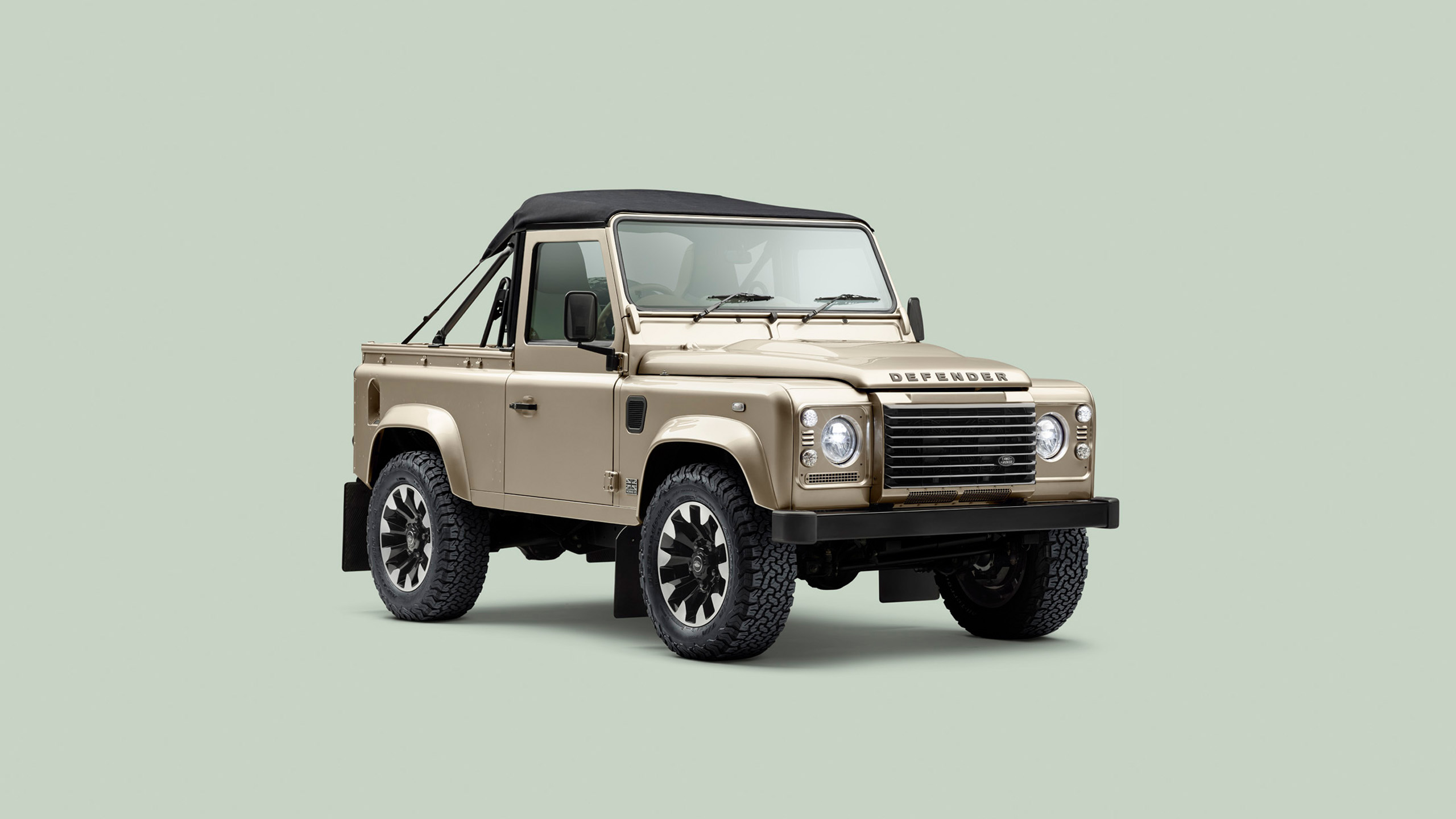 A beige Land Rover Defender convertible with a black soft top, rugged tires, and classic design, displayed against a plain light gray background.