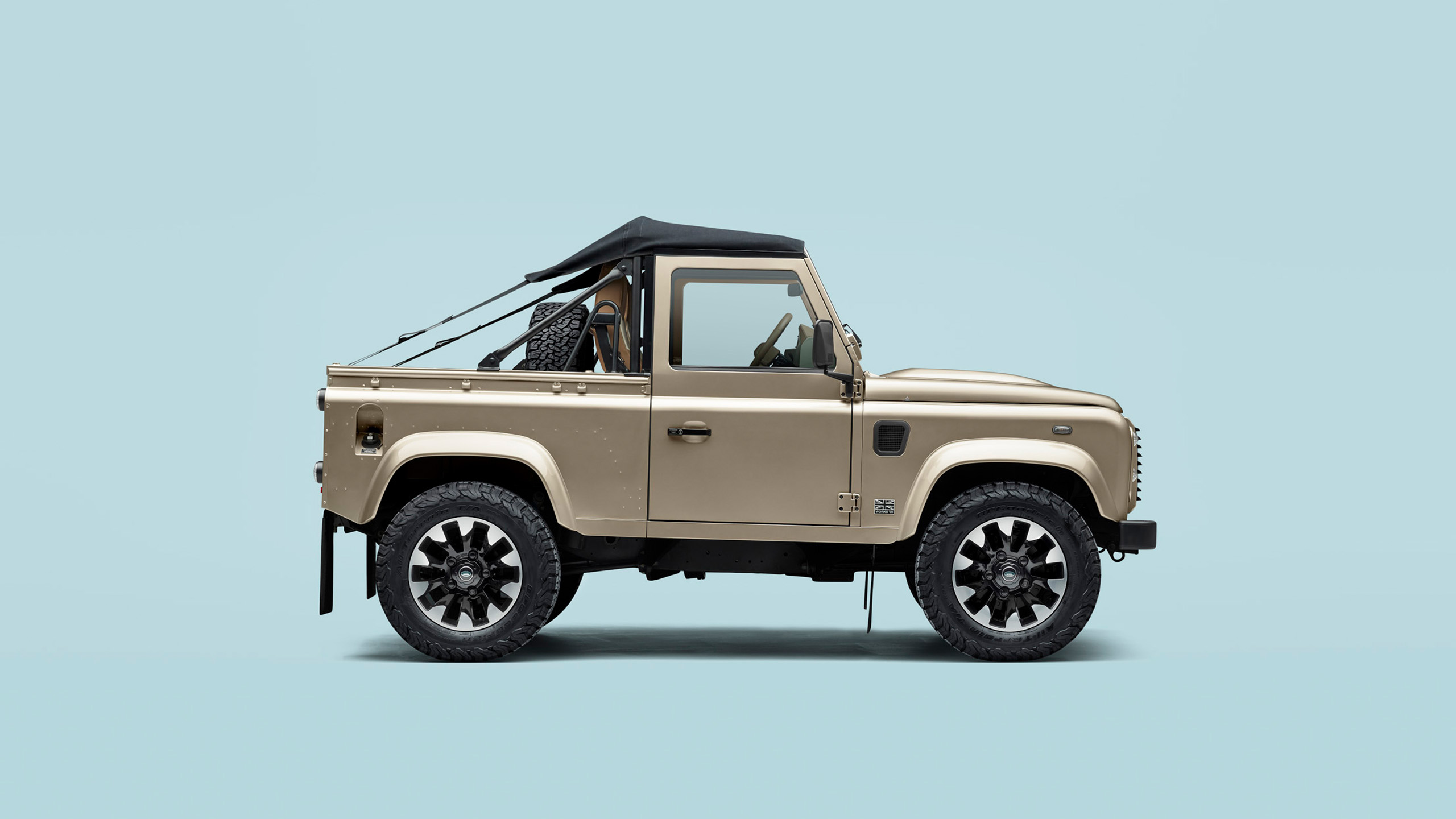 A beige, two-door Land Rover Defender with a black soft-top roof, viewed from the side against a plain light blue background.