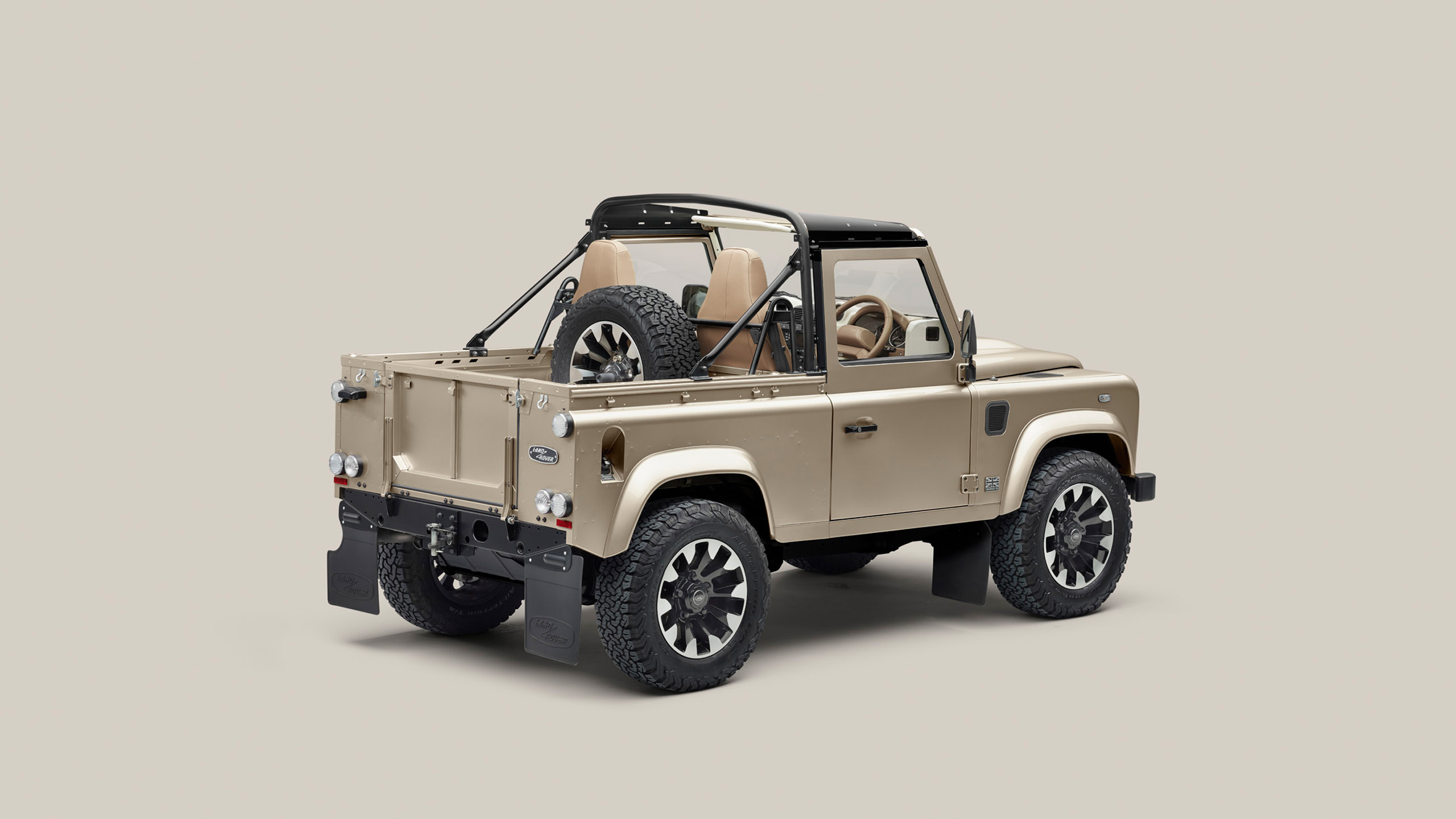 A beige, two-door off-road vehicle with an open cargo bed, roll bar, exposed spare tire behind the seats, and rugged tires, viewed from the rear left against a plain background.