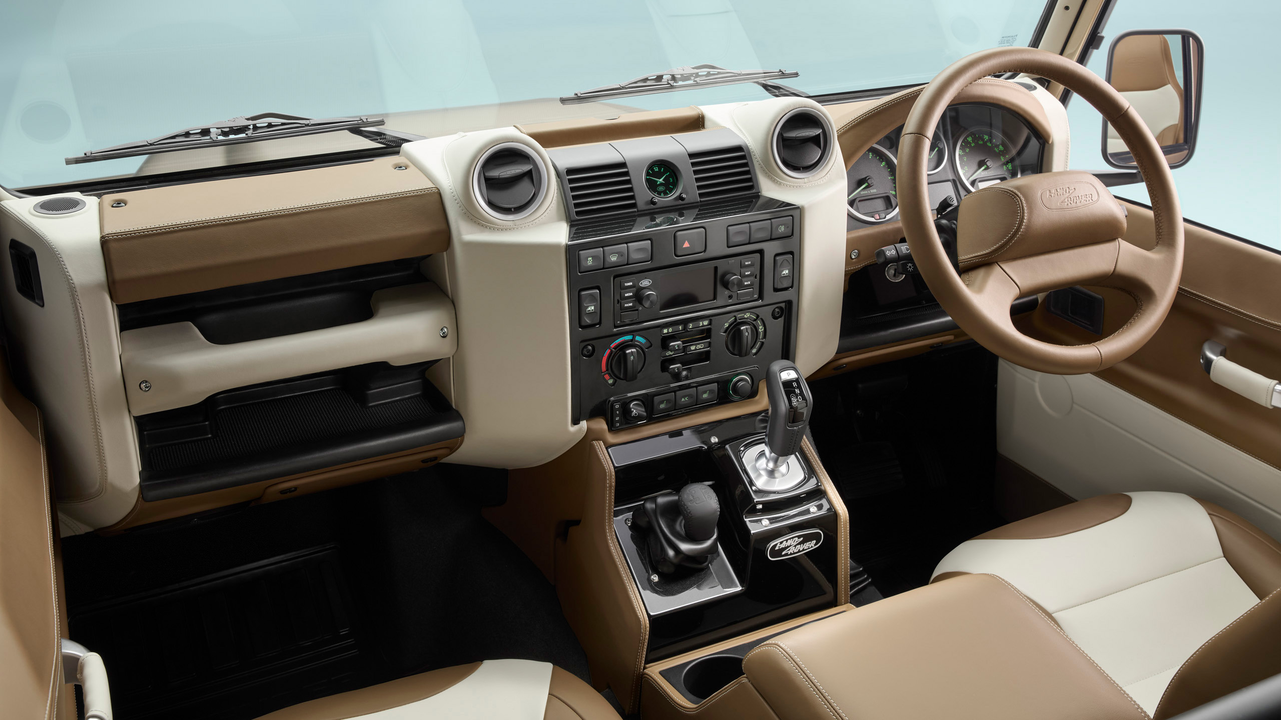 The image shows the interior of a Land Rover vehicle, featuring a brown and cream dashboard, leather seats, a manual gear shifter, and various control knobs and dials on the center console.