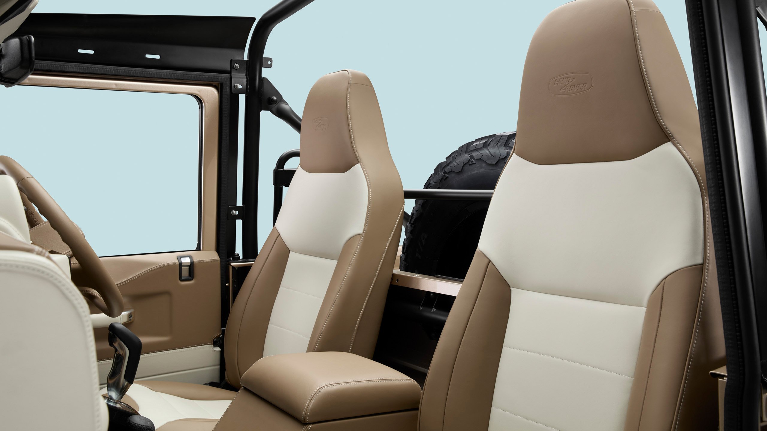Close-up view of two tan and cream leather car seats inside a rugged vehicle, with part of the door, a roll bar, and a spare tire visible in the background against a light blue backdrop.