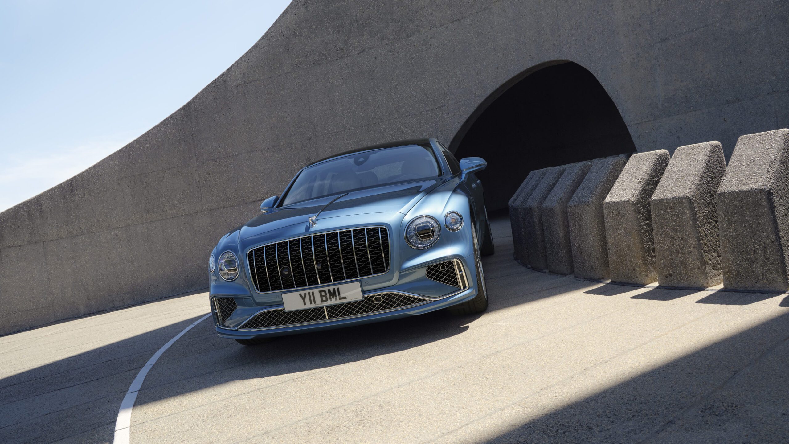 A blue luxury Bentley car is parked on a curved concrete road near a modern architectural tunnel, with sunlight casting shadows on the ground.