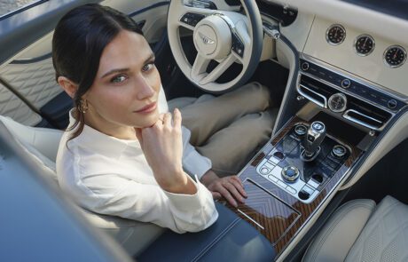 A woman in a white blouse sits in the driver’s seat of a luxury car, looking up toward the camera. The car has cream leather seats, wood paneling, and a modern dashboard with elegant controls.