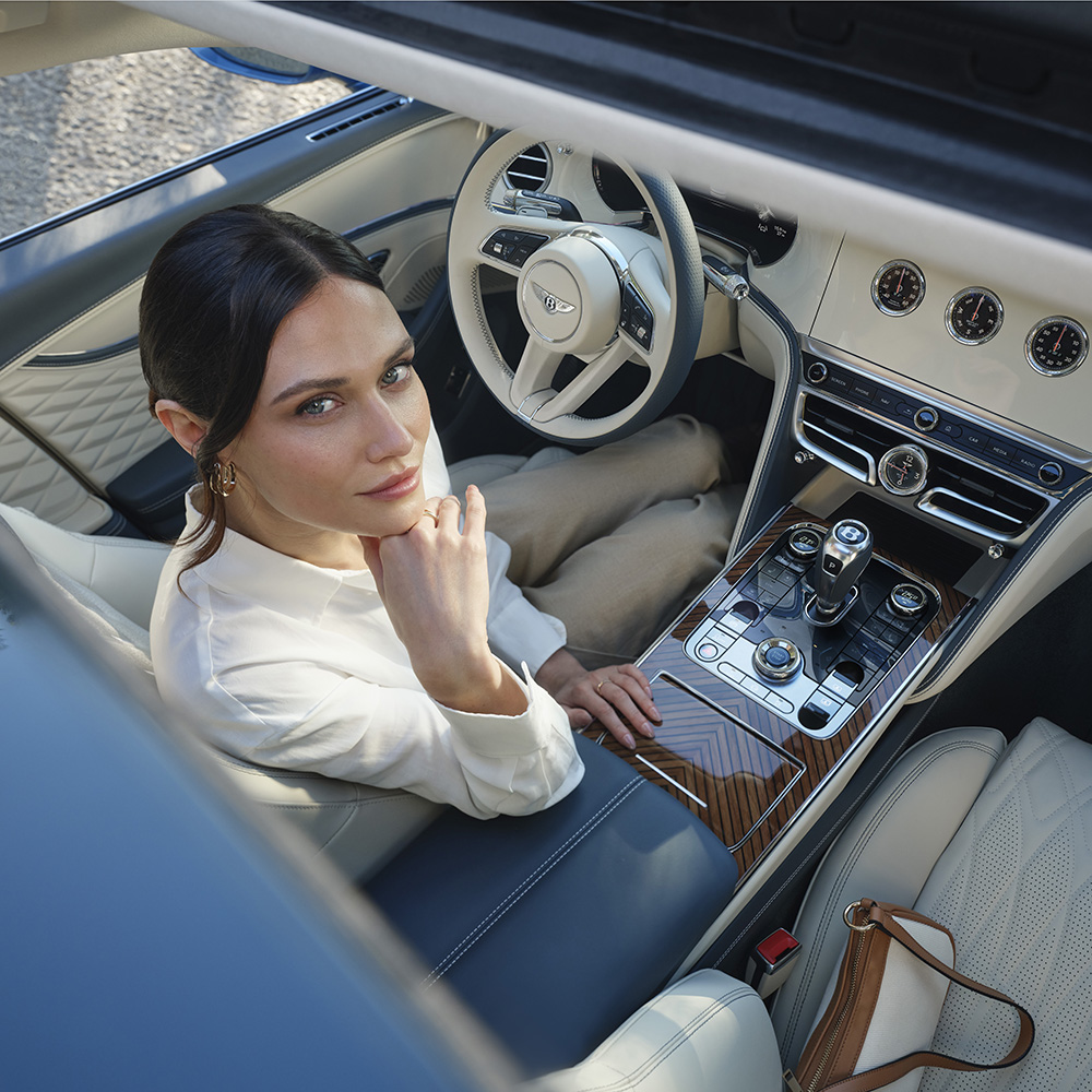 A woman in a white blouse sits in the driver’s seat of a luxury car, looking up toward the camera. The car has cream leather seats, wood paneling, and a modern dashboard with elegant controls.