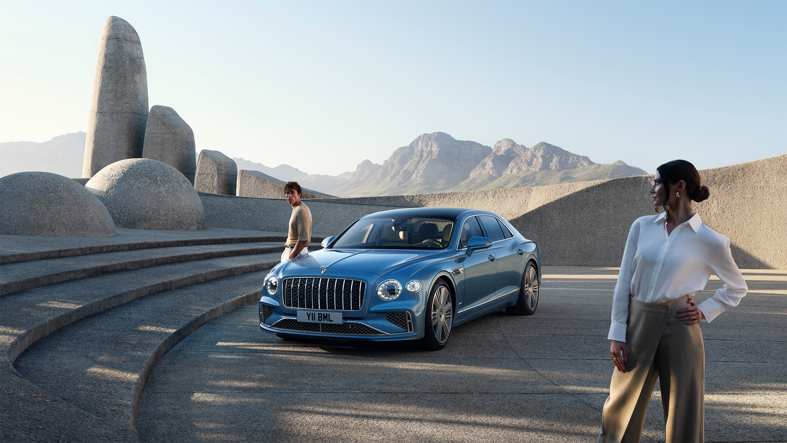 A blue luxury sedan is parked on a modern stone plaza with abstract sculptures. A man stands near the car and a woman stands in the foreground. Mountains and a clear sky are visible in the background.