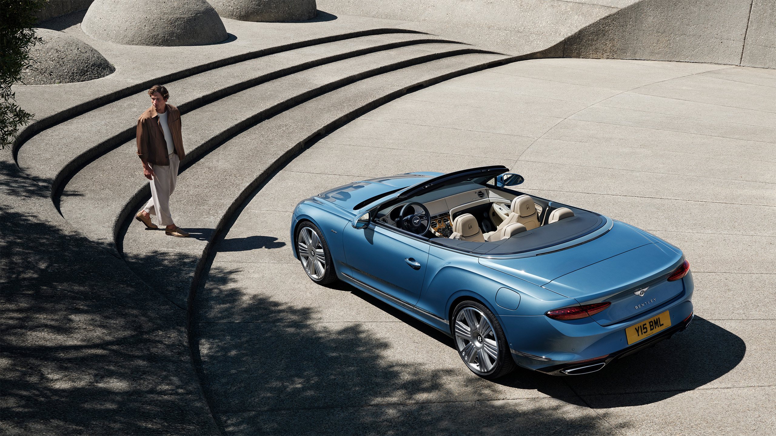 A person walks near curved concrete steps beside a blue convertible car with its top down, parked on a smooth, modern stone surface under sunlight.