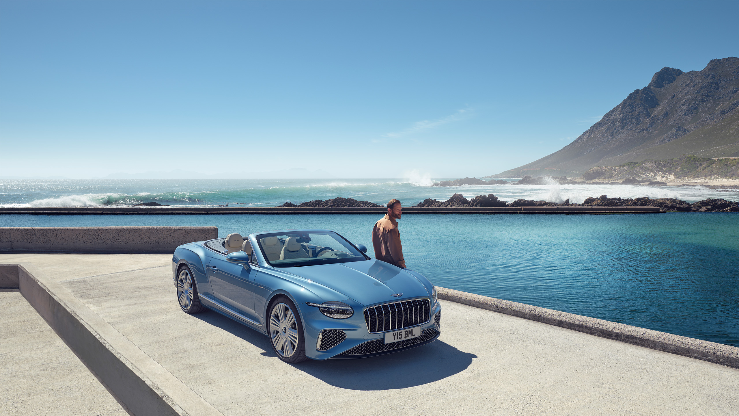 A blue convertible luxury car is parked near a pool by the ocean, with a man in swim trunks standing beside the water. Mountains and waves are visible in the background under a clear blue sky.