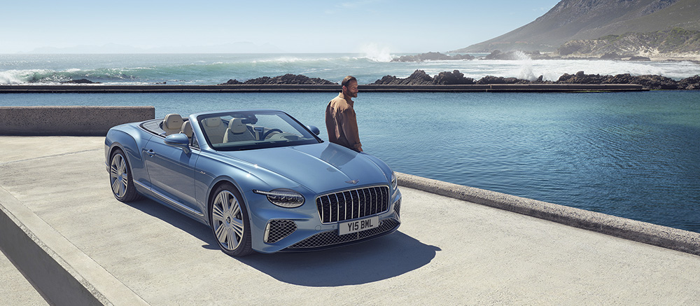 A blue luxury convertible car is parked by a scenic oceanfront, with a man standing nearby looking at the water and waves crashing against rocky shores under a clear sky.