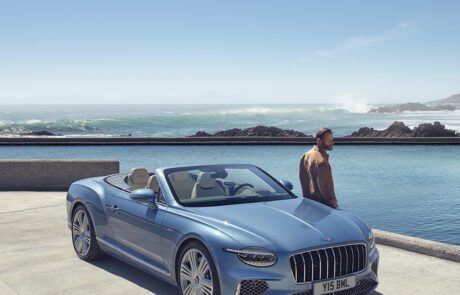 A man stands near a light blue convertible parked beside a pool with an ocean and rocky shoreline in the background under a clear sky.