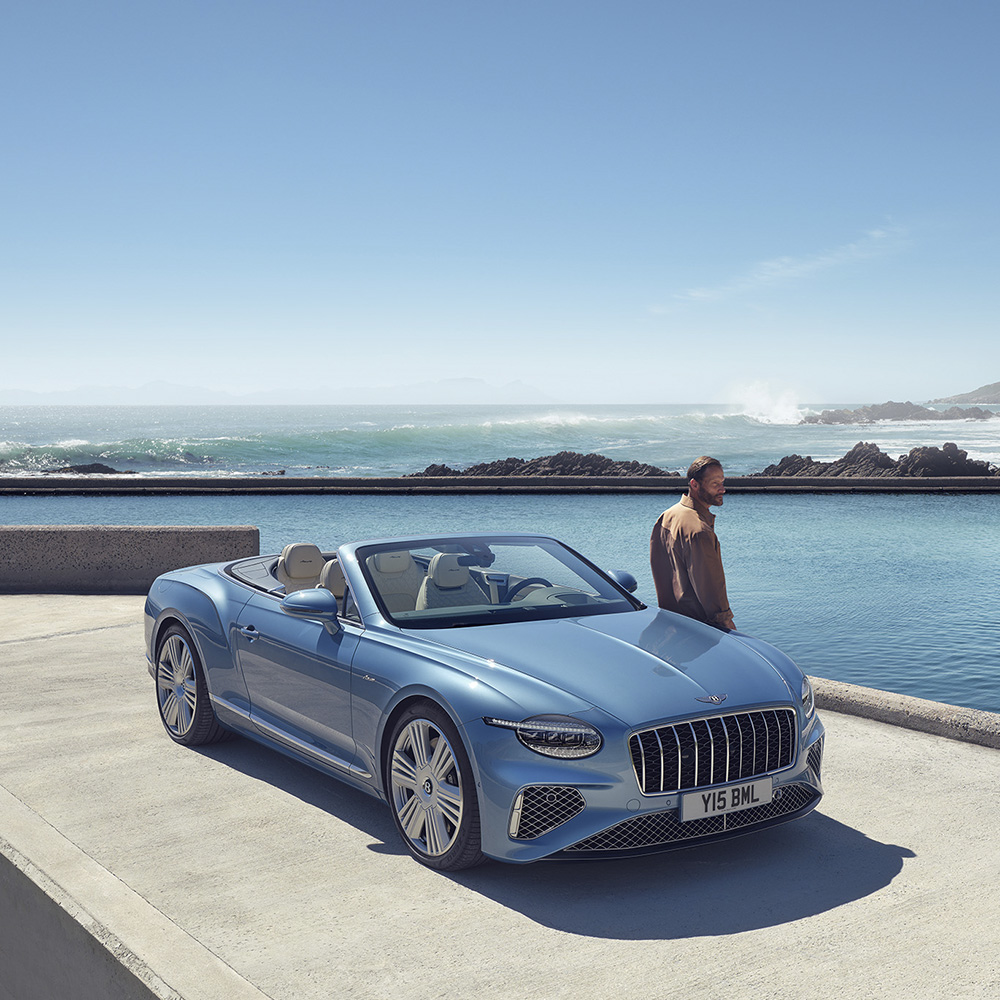 A man stands near a light blue convertible parked beside a pool with an ocean and rocky shoreline in the background under a clear sky.