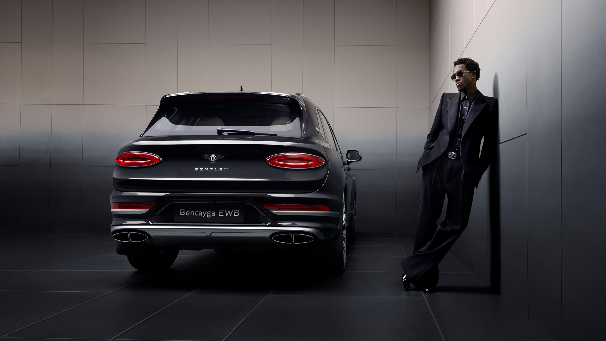 A person in a dark suit and sunglasses leans against a wall next to a black Bentley Bentayga EWB SUV, shown from the rear in a modern, minimalist room.