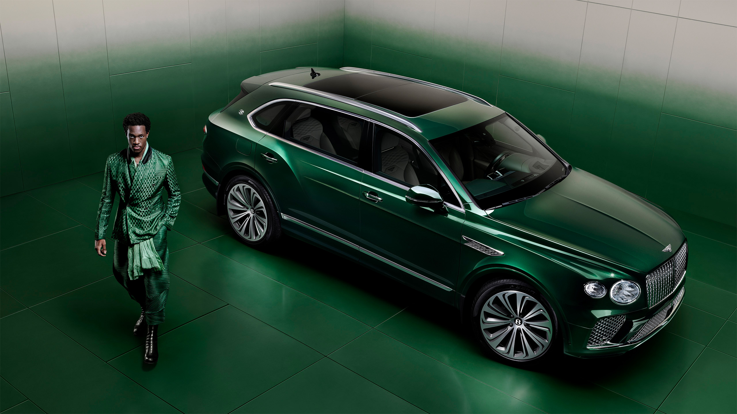 A man in a stylish green outfit walks past a luxury green Bentley SUV parked in a modern, green-toned room with an ombre wall and floor design.