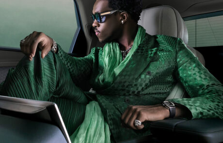 A stylish man in an emerald green suit and sunglasses relaxes in the luxurious back seat of a Bentley, gazing out the window. The interior features plush, light-colored upholstery and elegant details.