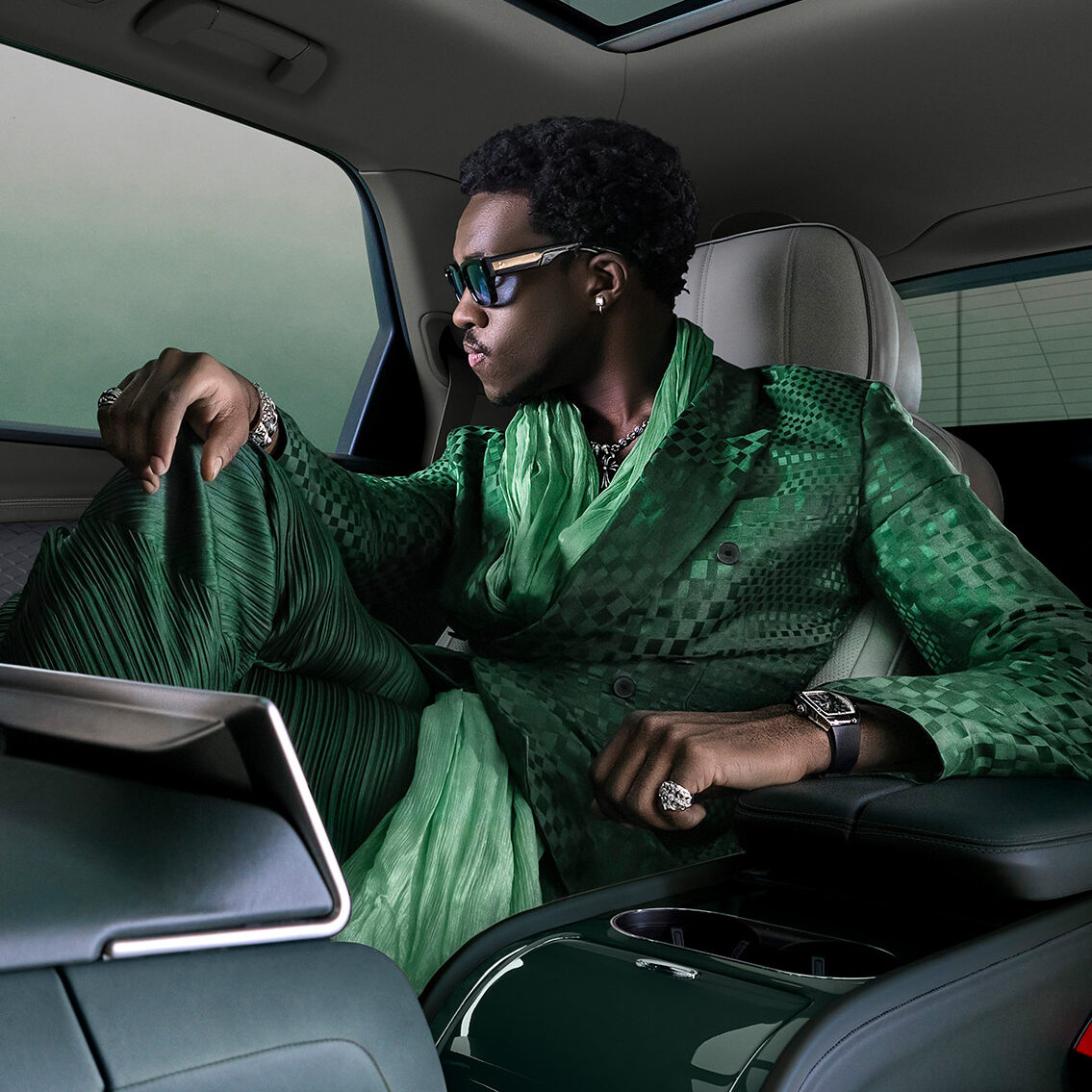 A stylish man in an emerald green suit and sunglasses relaxes in the luxurious back seat of a Bentley, gazing out the window. The interior features plush, light-colored upholstery and elegant details.