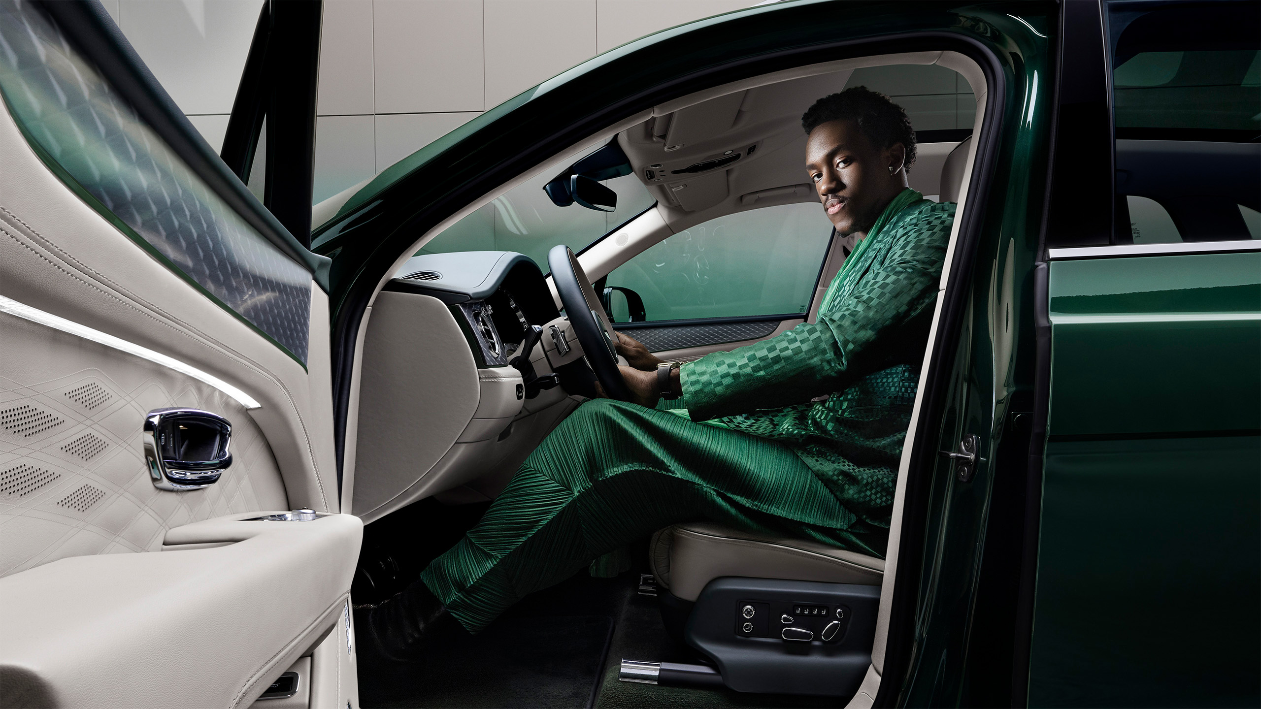 A person wearing a stylish green outfit sits in the driver’s seat of a luxury car with the driver’s door open, looking at the camera and smiling. The car interior is white and modern.