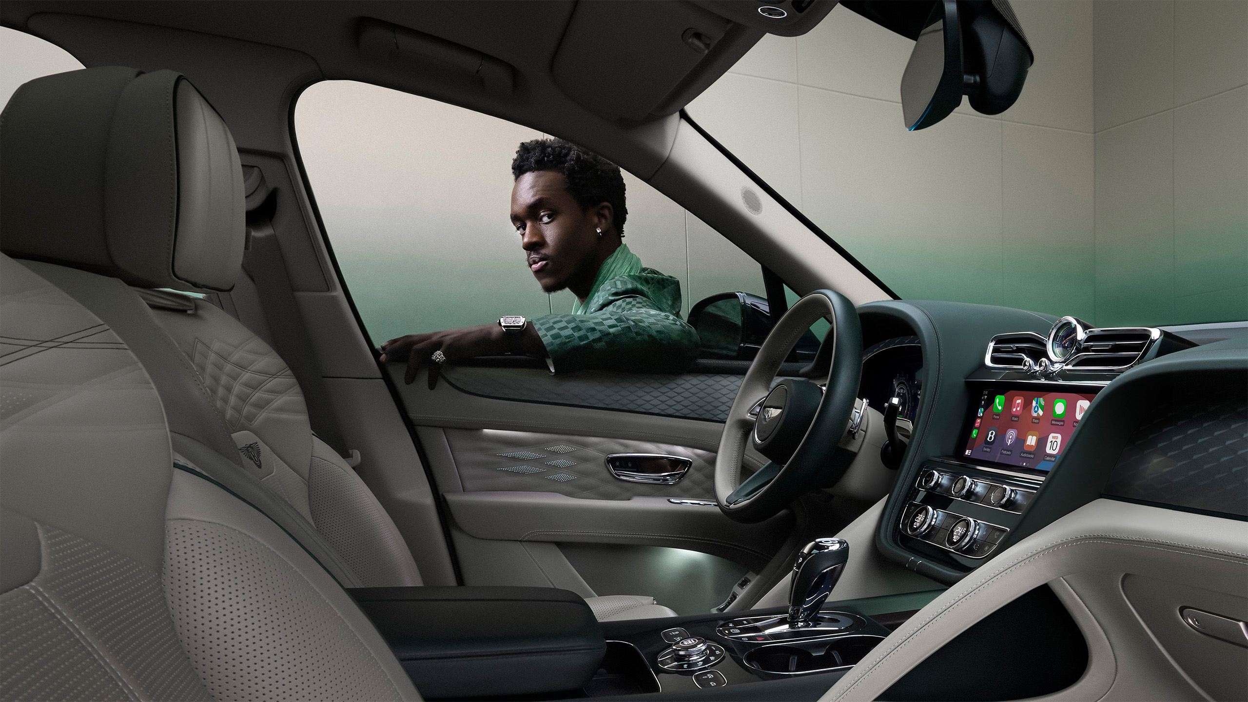 A man wearing a green jacket leans on the open driver’s side window of a modern luxury car, seen from the perspective of the front passenger seat. The car’s dashboard and infotainment system are visible.