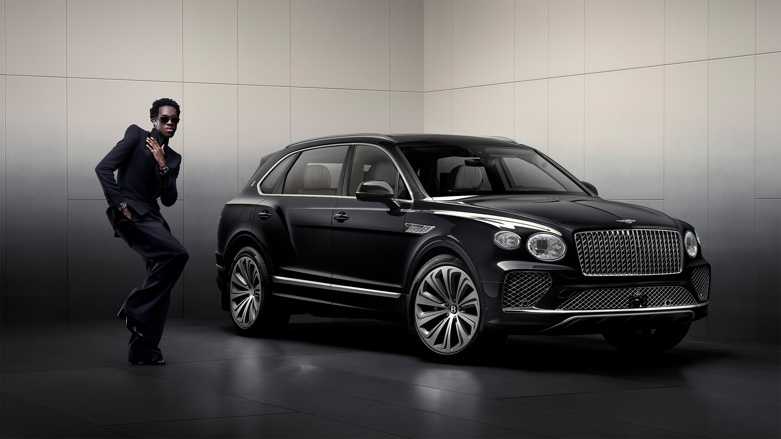 A man in a black suit poses confidently next to a sleek black luxury SUV in a modern, minimalist showroom with neutral-colored walls.