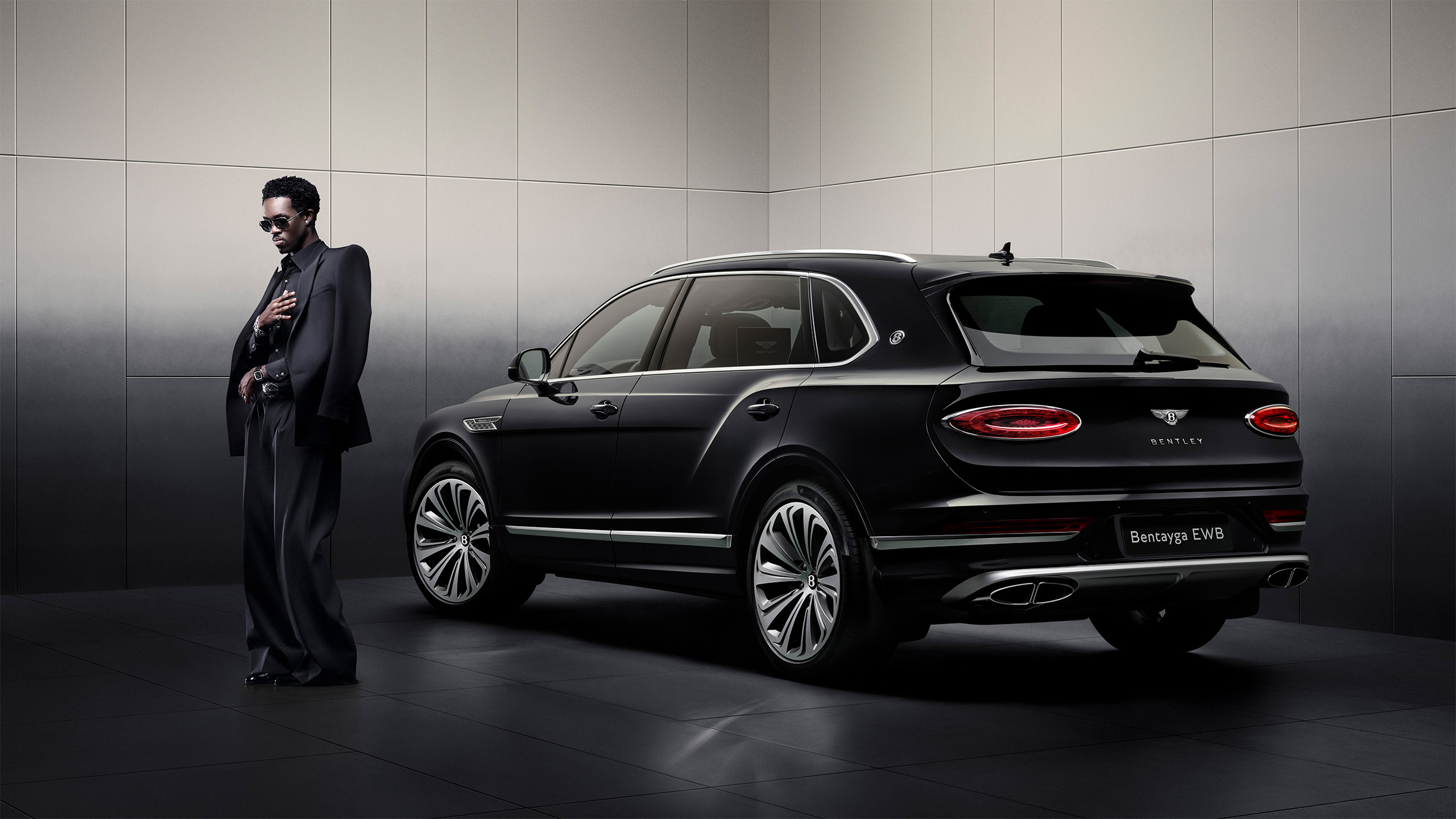 A man in a dark suit and sunglasses stands next to a sleek black Bentley Bentayga EWB SUV in a modern, minimalist showroom with soft lighting.