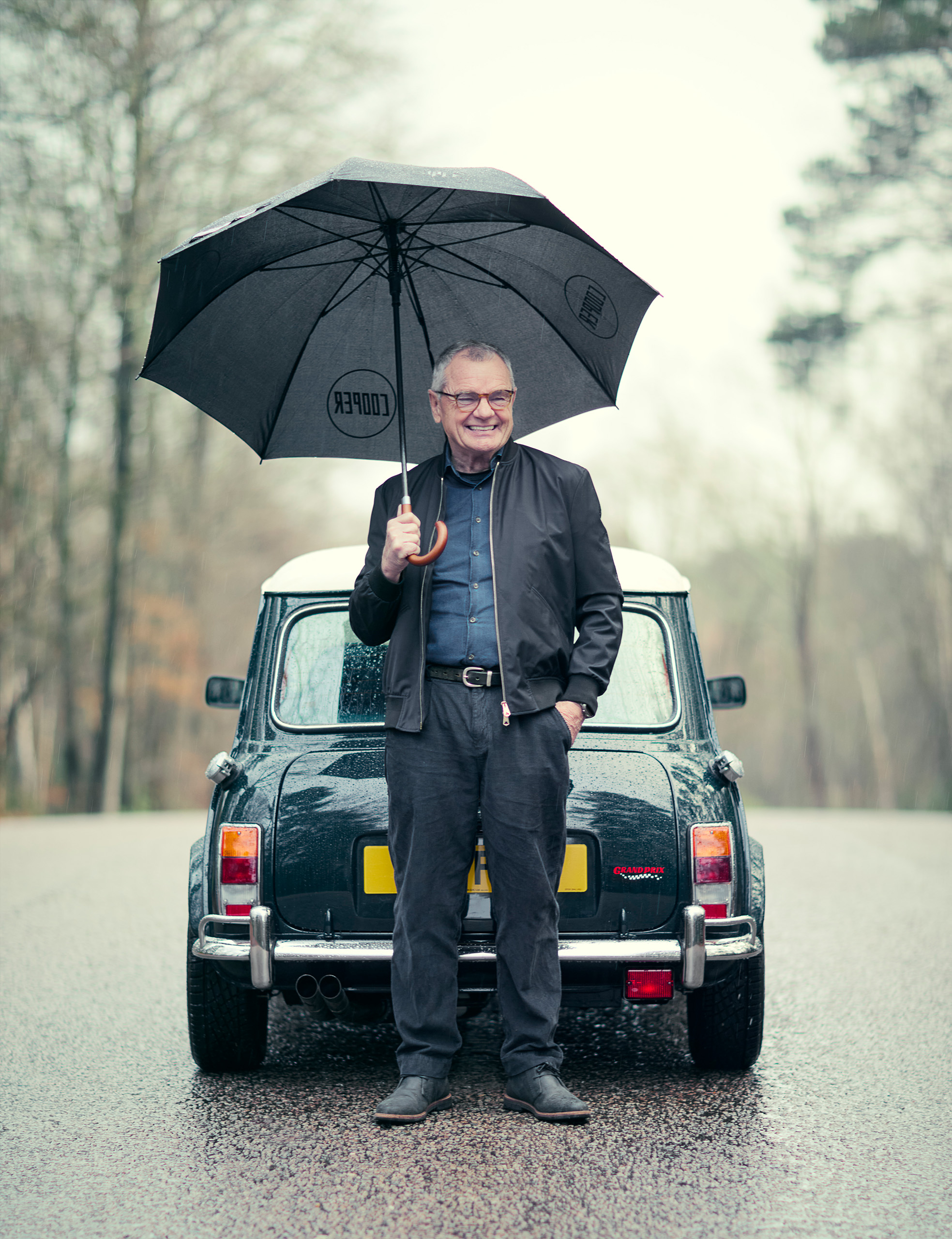 mike-cooper-obe-mini-cooper-grand-prix-umbrella Mike Cooper OBE stands smiling under a black umbrella behind a British Racing Green Mini Cooper Grand Prix in the rain.