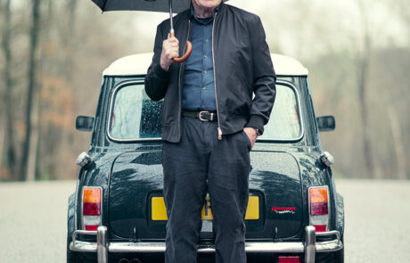 An older man smiles whilst standing in front of a classic Mini Cooper car on a rainy day, holding an open umbrella. The background shows a blurry, tree-lined road.