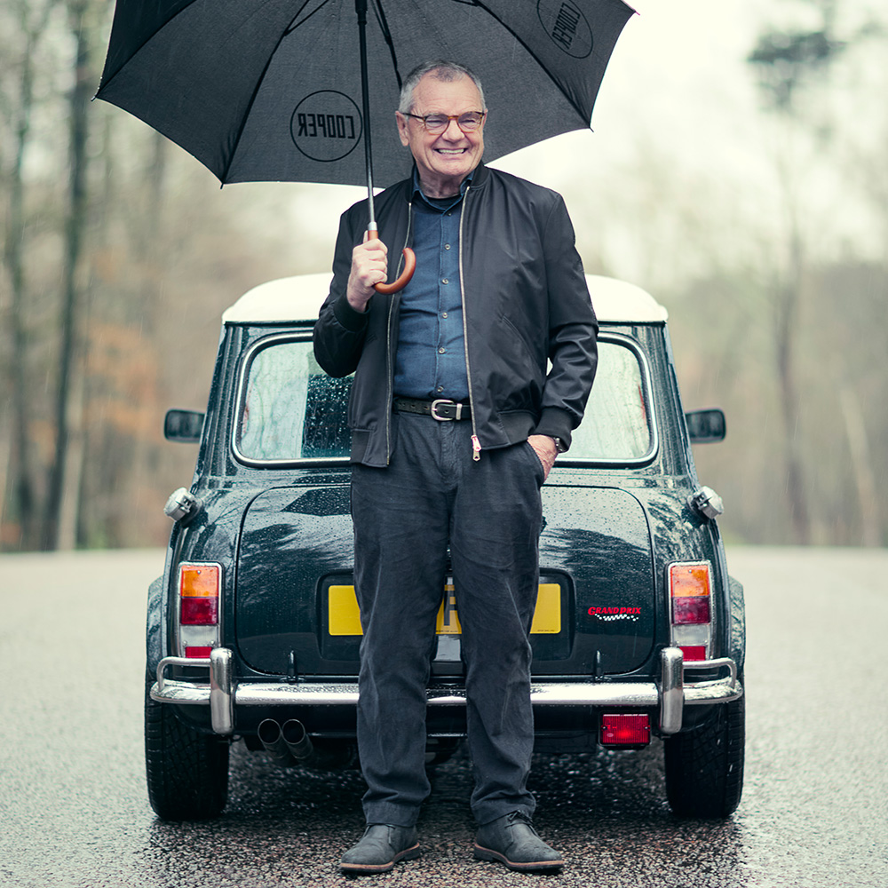 An older man smiles whilst standing in front of a classic Mini Cooper car on a rainy day, holding an open umbrella. The background shows a blurry, tree-lined road.