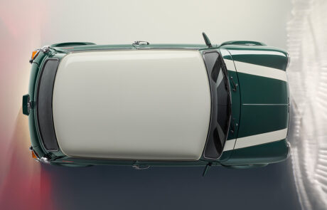 A top-down view of a green classic car with white racing stripes and a white roof, parked on a light-coloured surface with a gradient shadow to the side.