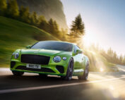 A bright green Bentley GT sports car drives on a winding mountain road with trees and sunlight in the background. The sun creates lens flare.