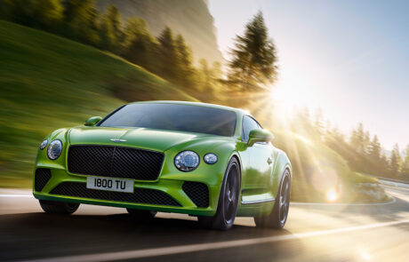 A bright green Bentley GT sports car drives on a winding mountain road with trees and sunlight in the background. The sun creates lens flare.