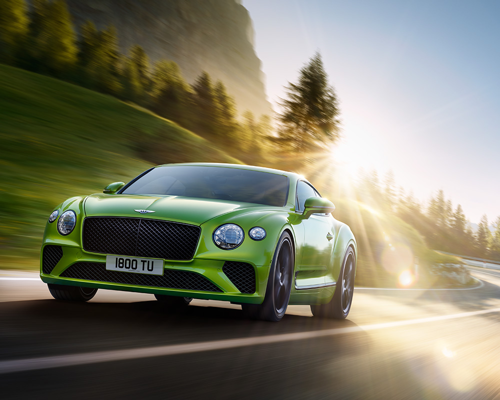 A bright green sports car drives on a winding mountain road with trees and sunlight in the background. The sun creates lens flare, and the car’s number plate reads “1800 TU”.