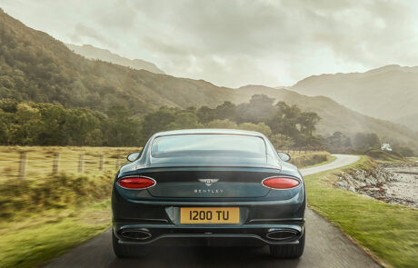 A dark green Bentley car drives on a narrow country road surrounded by grass, hills, and trees under a cloudy sky, with mountains visible in the distance.