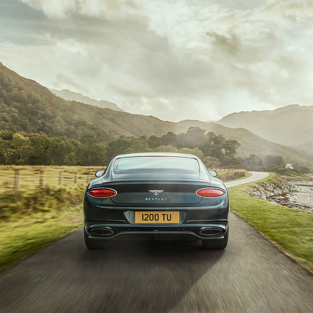 A dark green Bentley car drives on a narrow country road surrounded by grass, hills, and trees under a cloudy sky, with mountains visible in the distance.