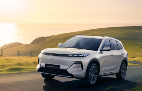 A white SUV drives on a scenic coastal road at sunset, with green hills and the sea in the background.