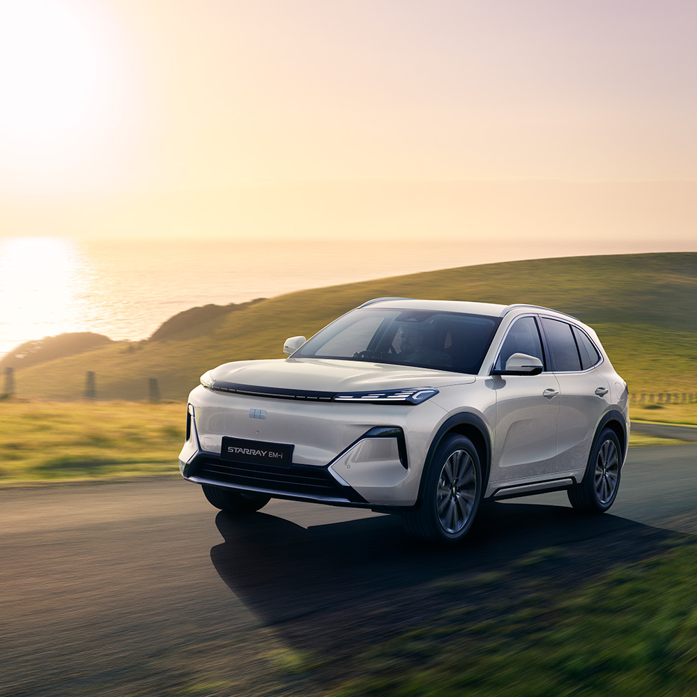 A white SUV drives on a scenic coastal road at sunset, with green hills and the sea in the background.