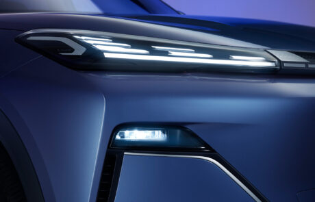 Close-up of a modern electric car’s front left headlamp and sleek blue body, with bright LED lights illuminated against a blue and purple gradient background.