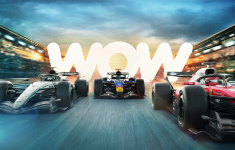 Three Formula 1 cars race side by side on a track at dusk, with glowing “WOW” text in the background, blurred stadium lights, and a dynamic sense of speed.