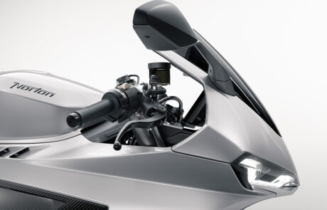Close-up of a sleek, silver Norton motorbike showcasing its angular wing mirror, handlebars, and modern headlamp against a plain, light background.