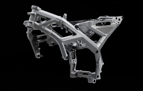 A metallic motorbike frame is suspended against a black background, showcasing its intricate structure and angular supports.