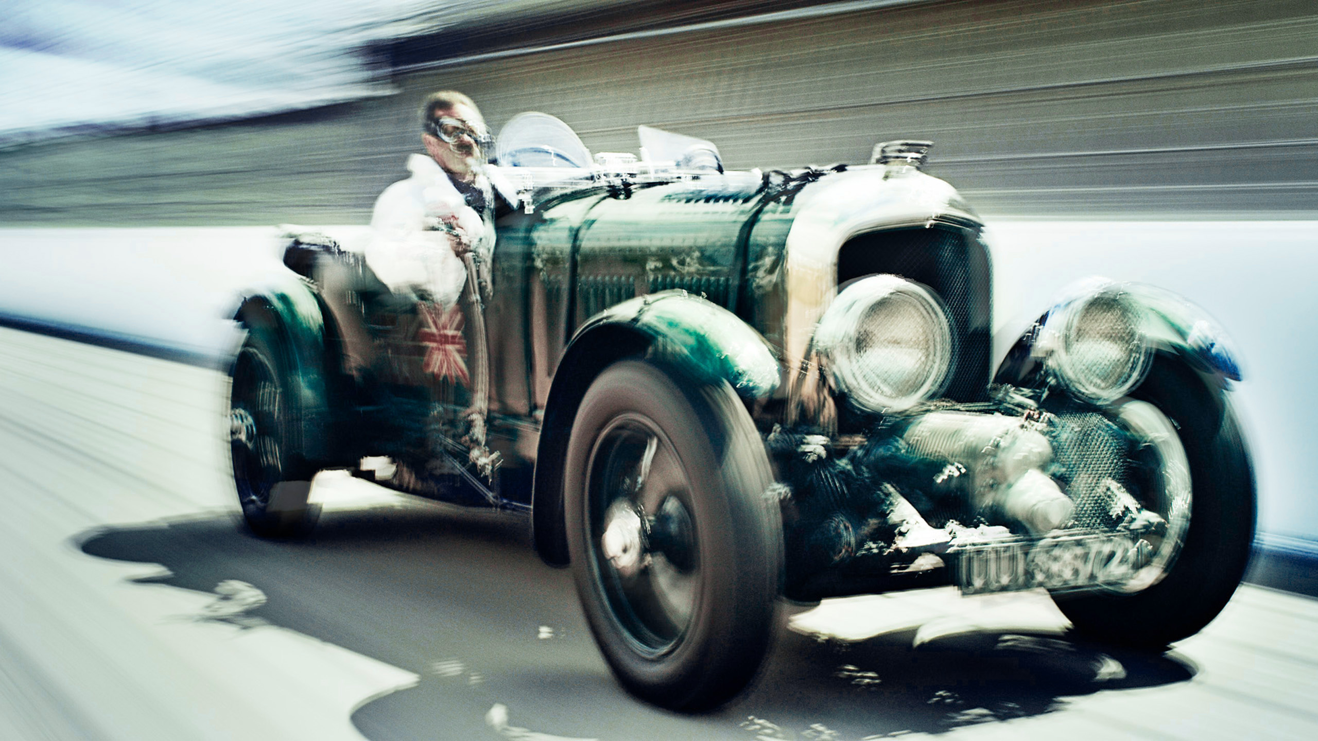 bentley-blower-vintage-le-mans-driver-front-quarter A vintage Bentley Blower racing car driven at speed by a driver in white overalls and cap, Union Jack livery visible on the bodywork.