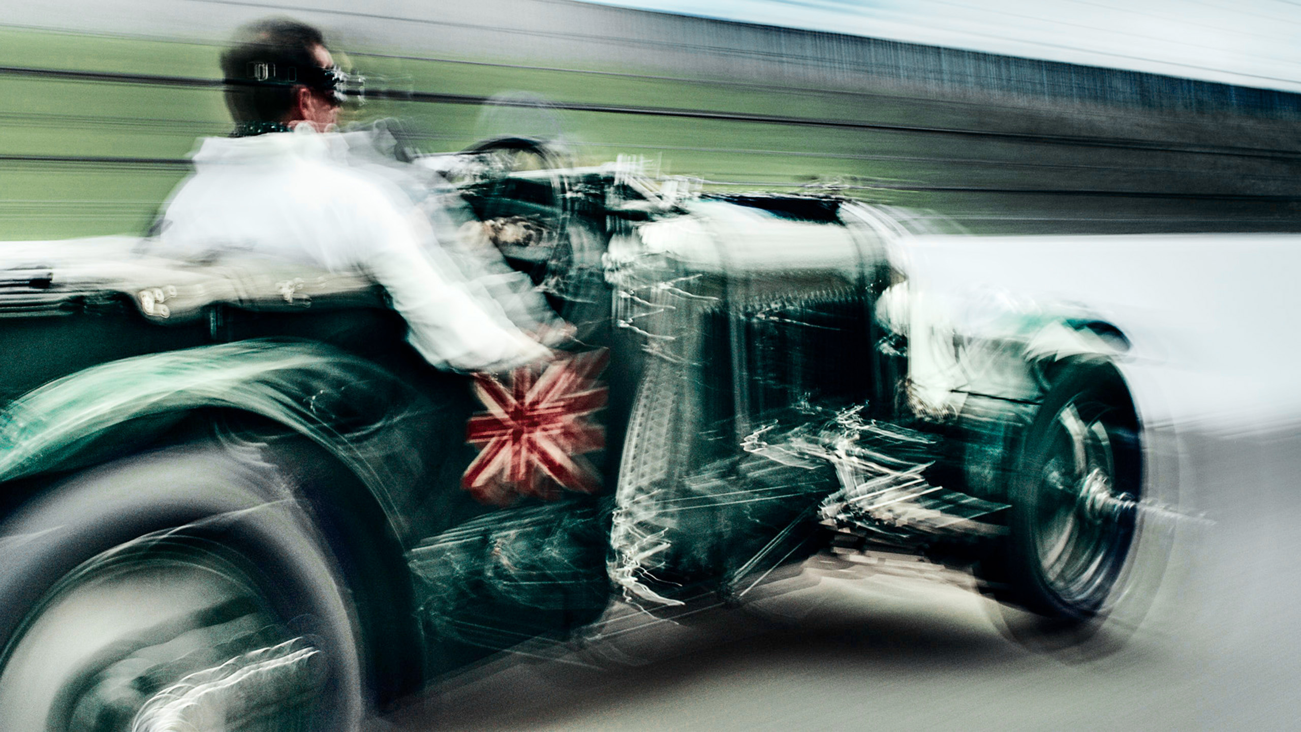 bentley-blower-vintage-le-mans-driver-side-profile Side view of a vintage Bentley Blower on track, the driver leaning into a corner with motion blur streaking the background.