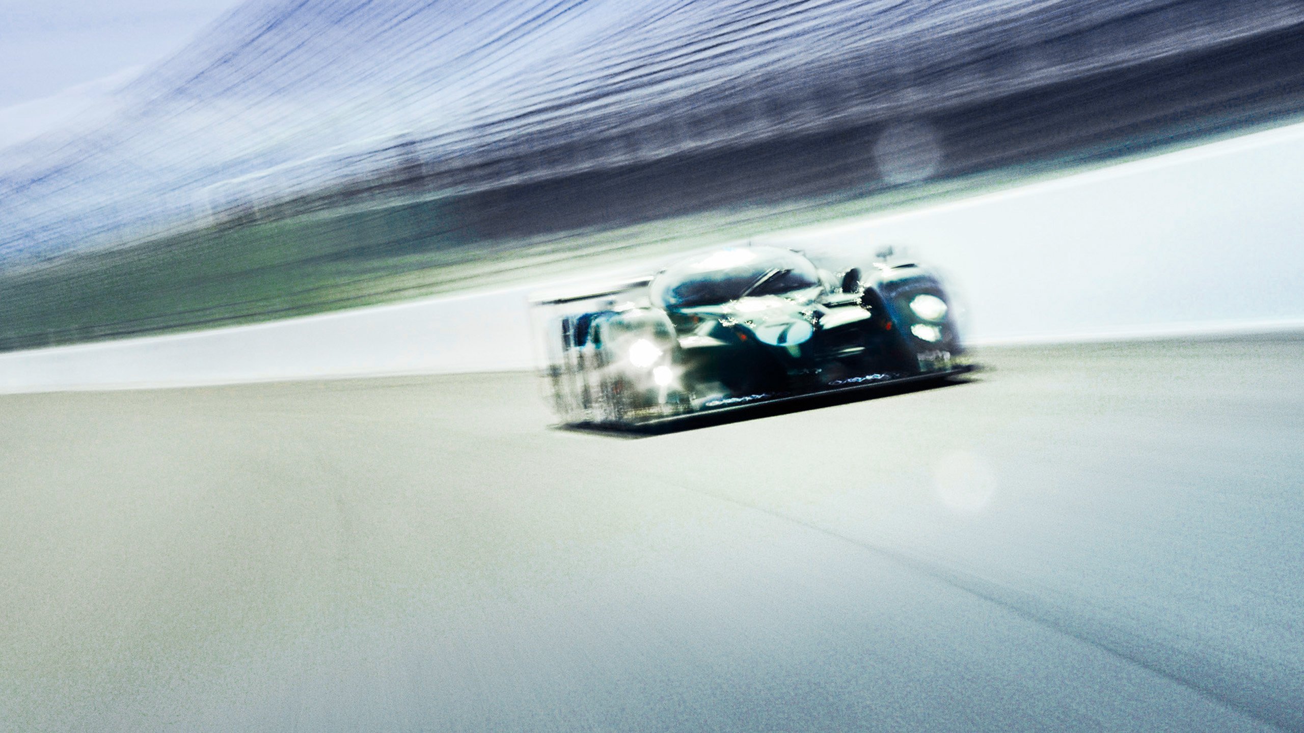 bentley-speed-8-le-mans-prototype-front-quarter-headlights A Bentley Speed 8 Le Mans prototype approaching head-on with headlights lit, heavy motion blur conveying high-speed pursuit on the racetrack.