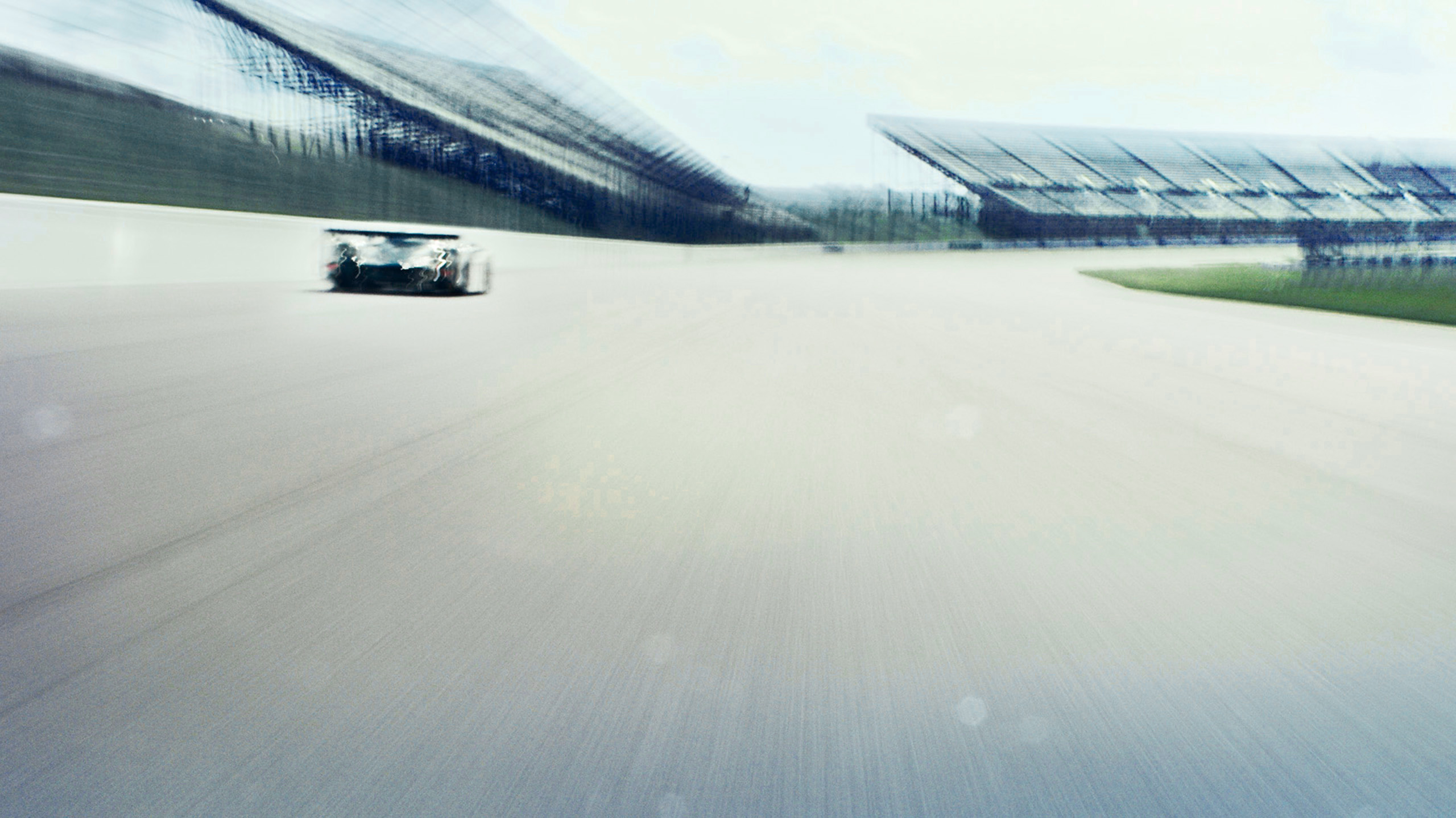 bentley-speed-8-le-mans-prototype-track-straight-wide A wide racetrack shot of a Bentley Speed 8 Le Mans prototype racing past grandstands, motion blur stretching the background into streaks.