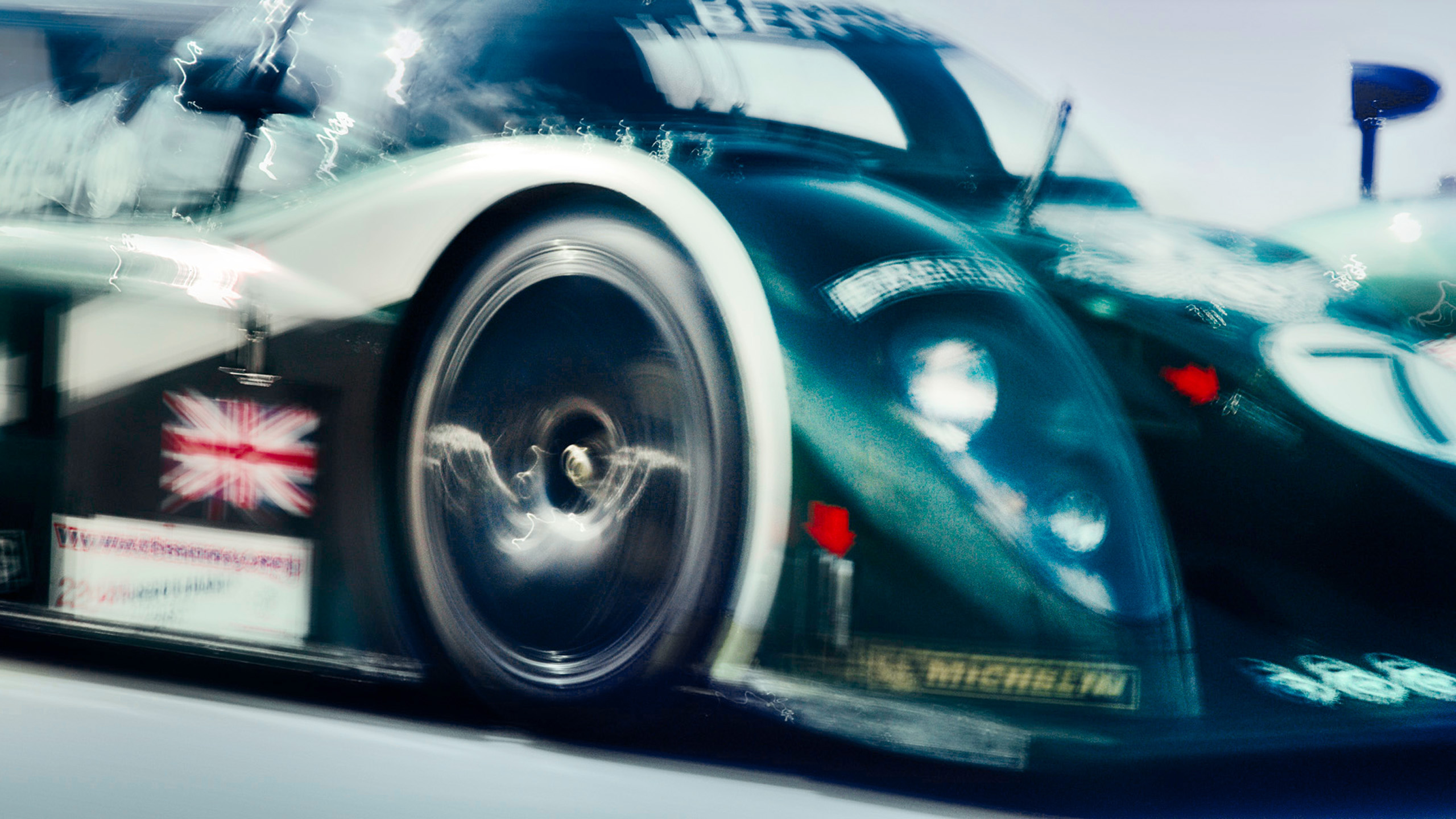 bentley-speed-8-le-mans-prototype-wheel-and-livery-detail Close-up of a Bentley Speed 8 Le Mans prototype's front wheel and Union Jack livery detail, captured with motion blur at racing speed.