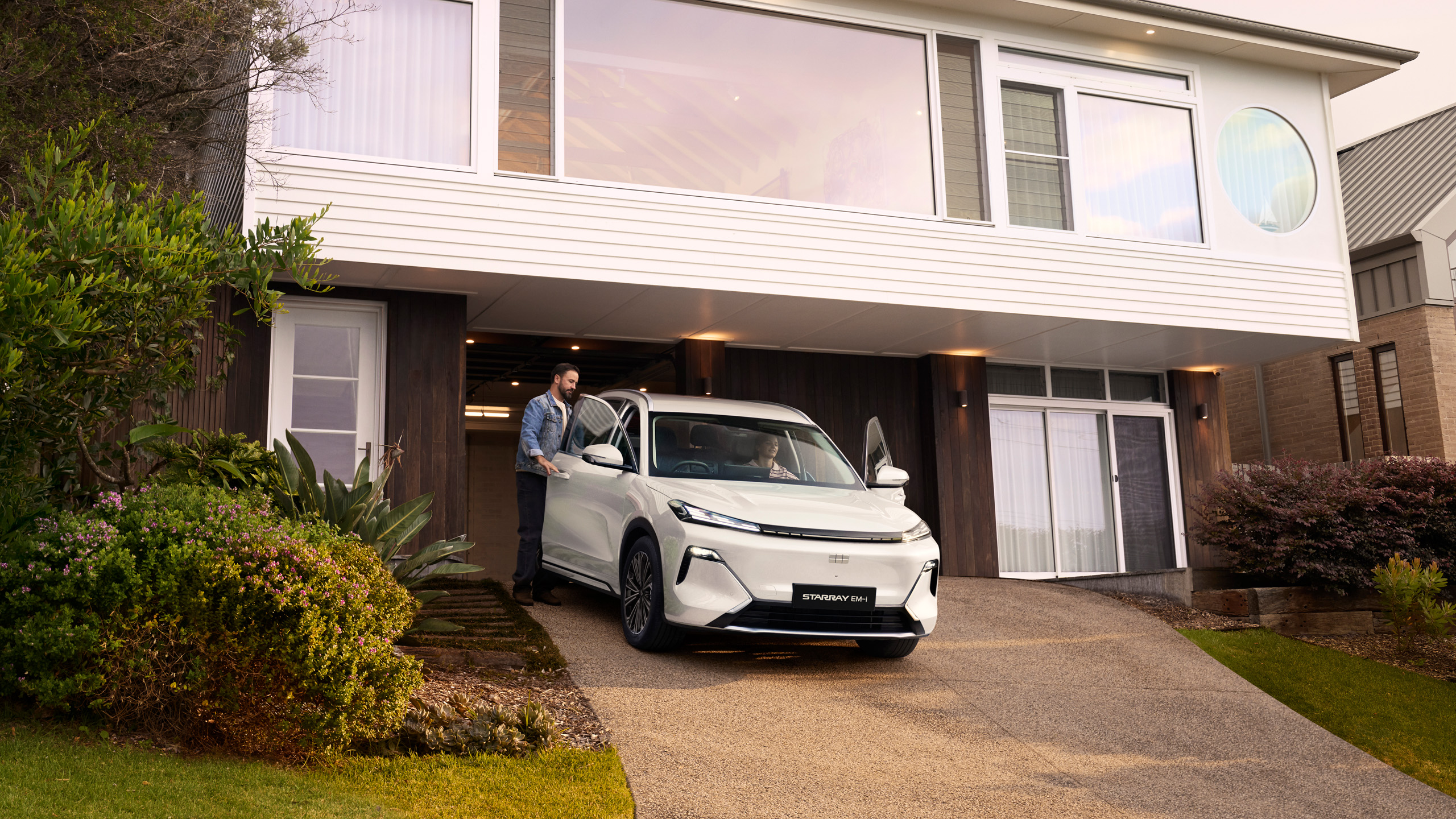 geely-starray-emi-leaving-home-dusk A man opens the door of the Geely Starray EM-i in a driveway at dusk, with a woman seated inside.