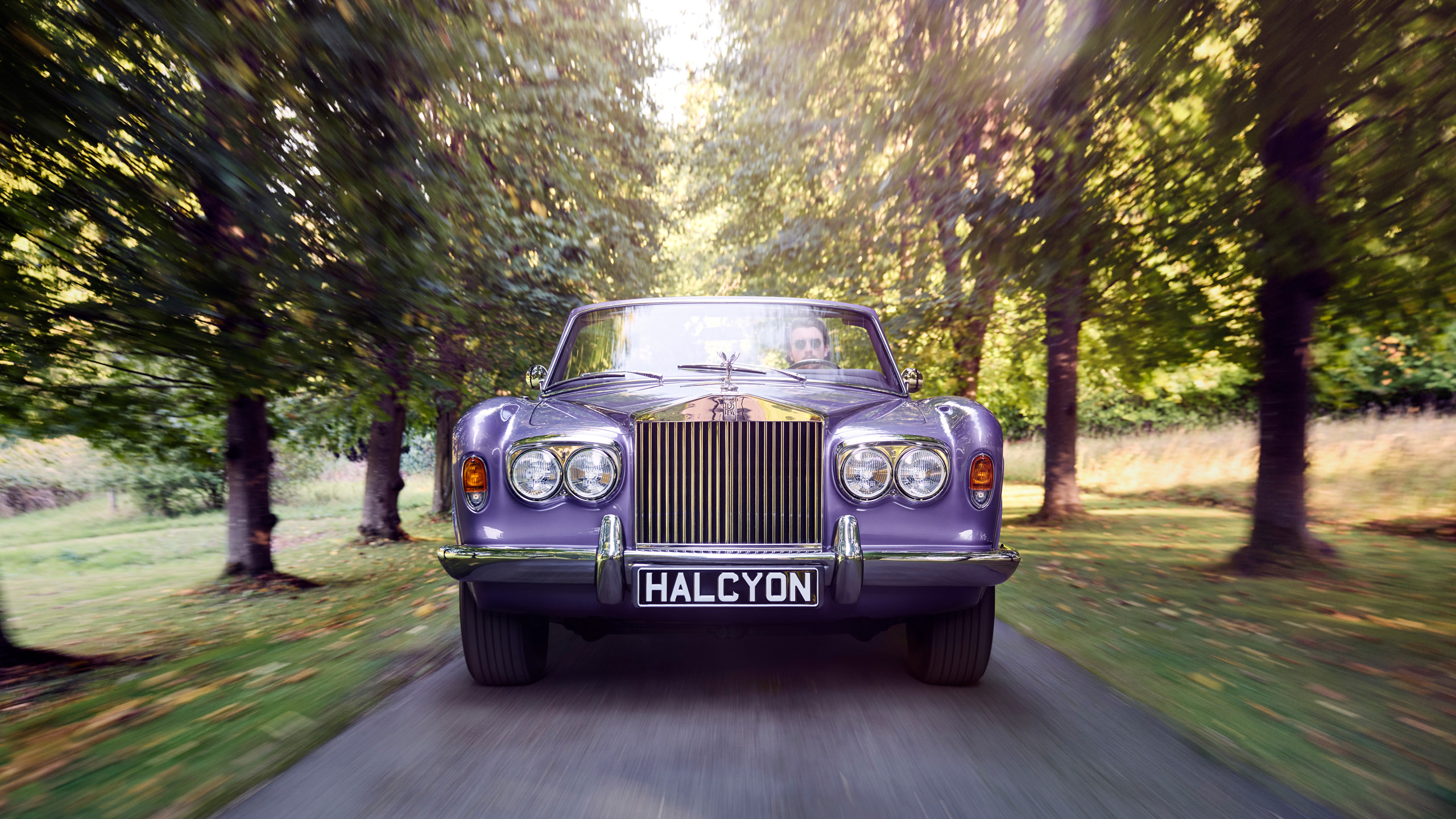 halcyon-electric-rolls-royce-corniche-driving-front-avenue The purple Halcyon electric Rolls-Royce Corniche driving toward camera along a tree-lined avenue, driver visible through the windscreen.