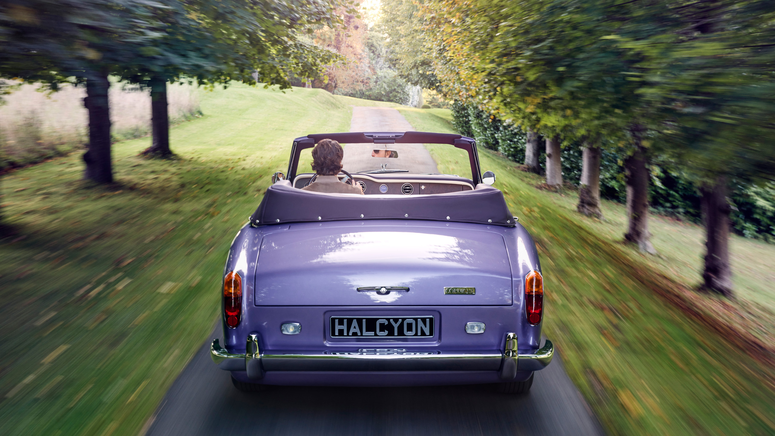 halcyon-electric-rolls-royce-corniche-driving-rear-avenue Rear view of the Halcyon electric Rolls-Royce Corniche driving away along a tree-lined avenue, with driver and passenger aboard.