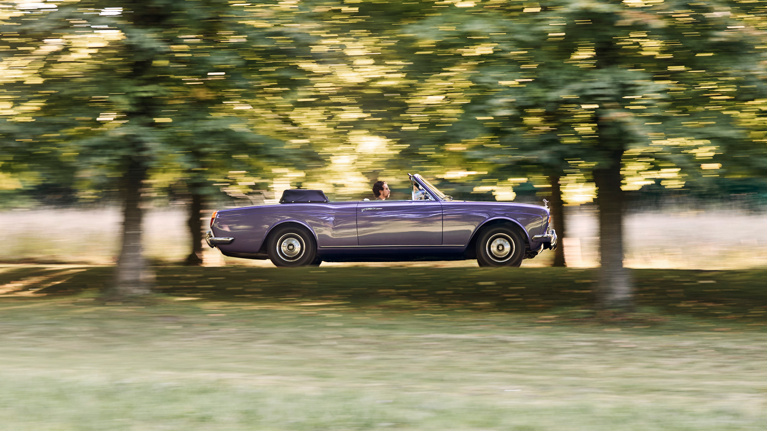 halcyon-electric-rolls-royce-corniche-driving-side-profile Side profile of the Halcyon electric Rolls-Royce Corniche driving along a tree-lined avenue, motion blur in the background.