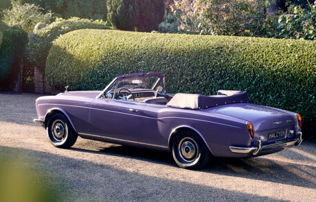 Square-format rear three-quarter view of the Halcyon electric Rolls-Royce Corniche parked beside clipped garden hedges.