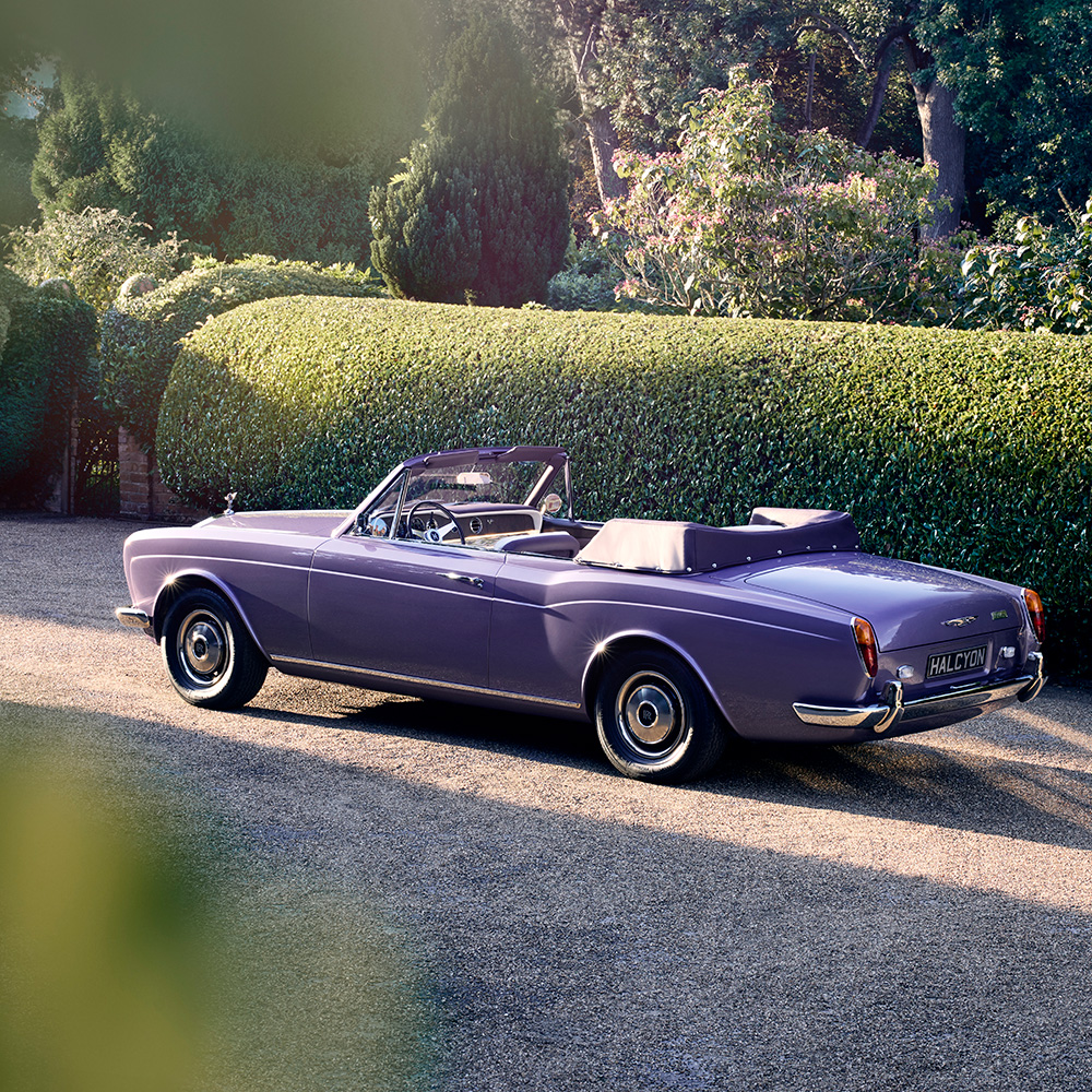 Square-format rear three-quarter view of the Halcyon electric Rolls-Royce Corniche parked beside clipped garden hedges.