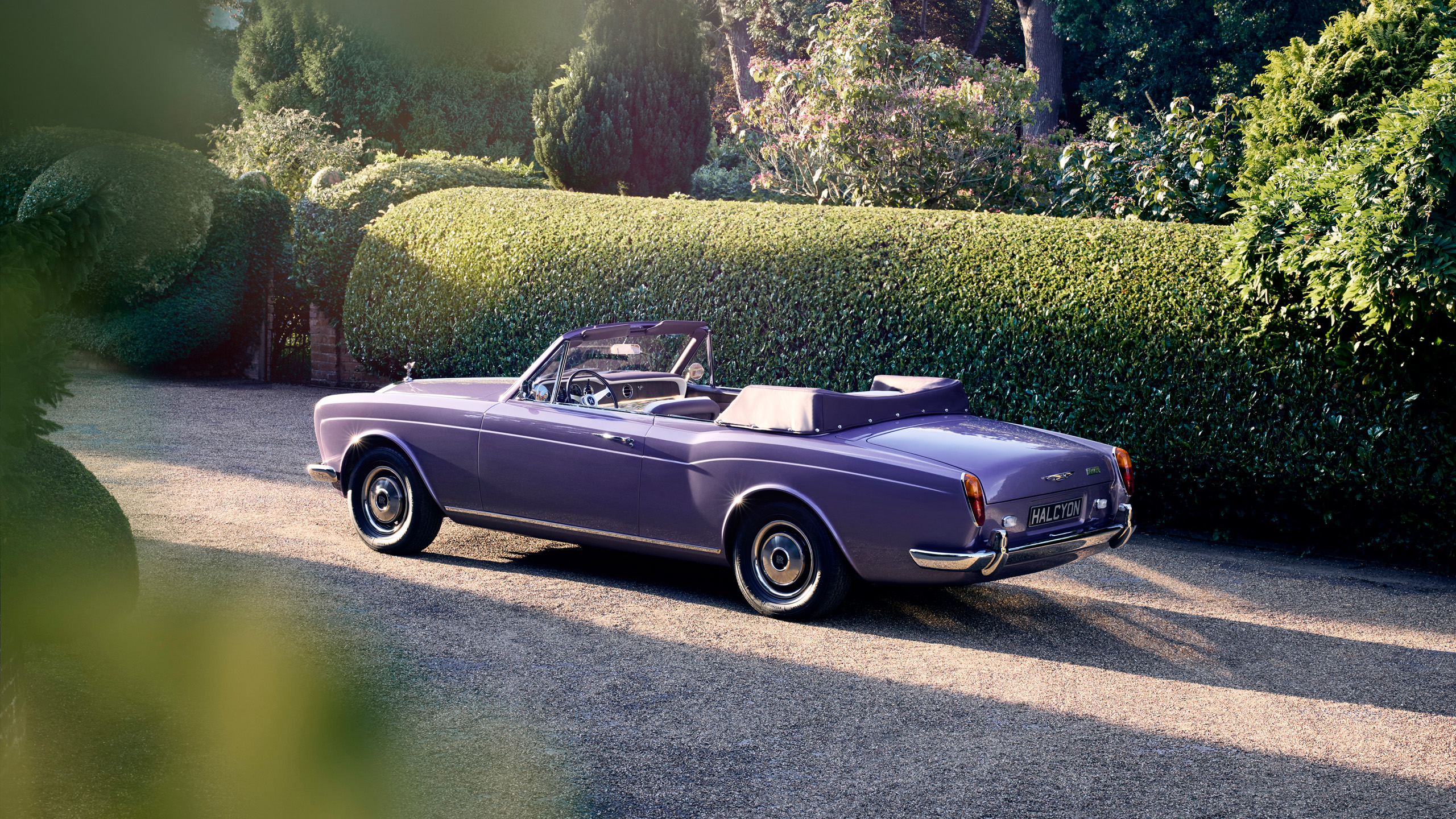 halcyon-electric-rolls-royce-corniche-rear-three-quarter-garden Rear three-quarter view of the Halcyon electric Rolls-Royce Corniche parked on a gravel driveway beside clipped hedges.