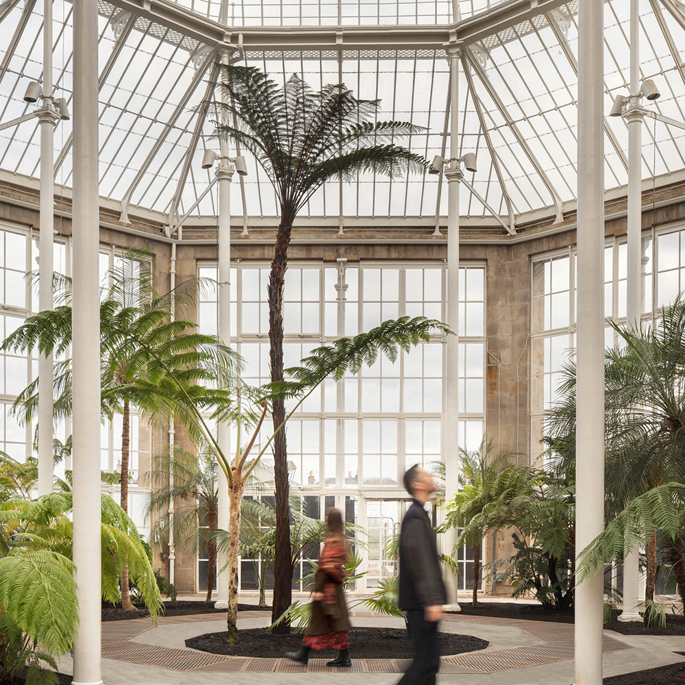 Two people walk inside a bright glass conservatory with tall palm trees and lush green plants. The structure features large windows and a geometric glass ceiling, letting in abundant natural light.